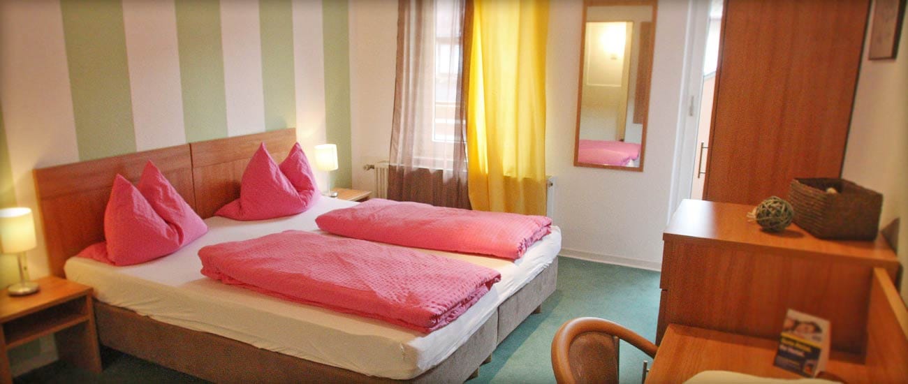 Doppelzimmer mit pinkfarbener Bettwäsche, grün-weiß gestreifter Wand, gelben Vorhängen und Holzmöbeln